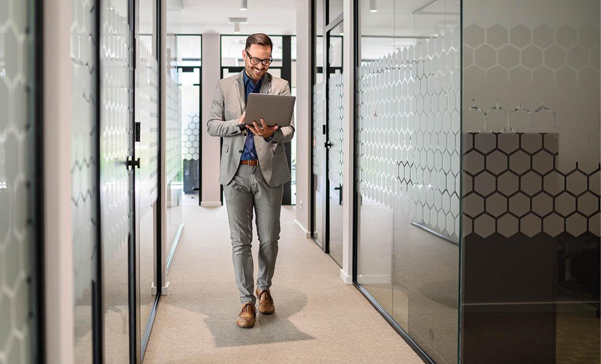 Full length of male software developer researching online over laptop while walking in office corridor
