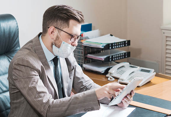 Young businessman in glasses and face mask working on a tablet in his personal office  Business during quarantine 