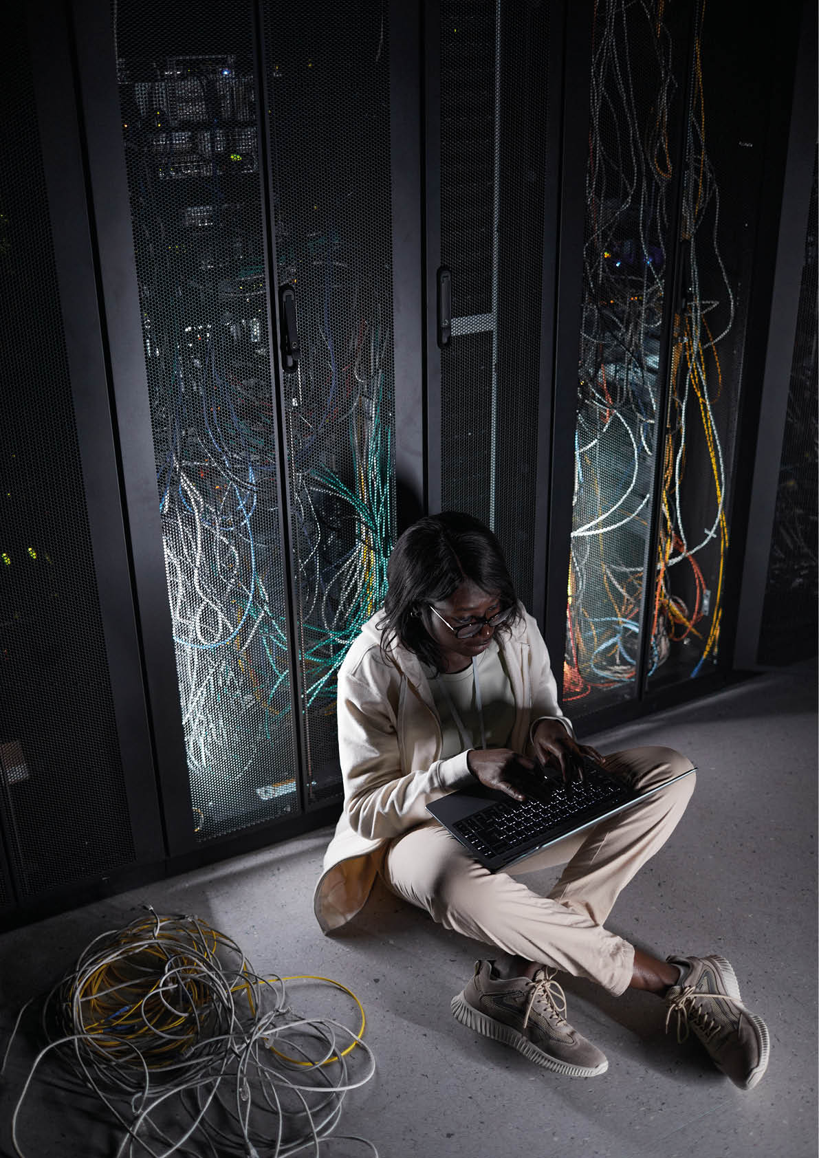 Vertical full length portrait of young African-American woman sitting on floor in server room and using laptop while working with supercomputer at data center, copy space