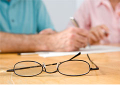 Dos personas mayores firmando un papel y unas gafas en la mesa