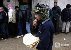 un niño come pan mientras varias personas esperan junto a una panadería en Al Qusayr, una ciudad del oeste de Siria