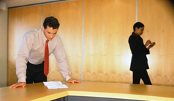 Un hombre de traje leyendo un documento mientras una mujer se pasea cerca suya por un despacho.