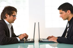 Dos ejecutivos frente a frente mirando a su portátil cada uno, sentados en una mesa.