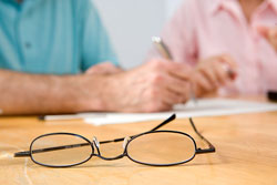 Unas gafas sobre una mesa en la que dos personas están escribiendo sobre un papel